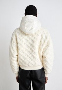 Krämfärgad texturerad tröja med en puffig vit huva, med ett kviltat mönster och ribbade muddar. I kombination med svarta läderbyxor.