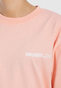 Lys rosa bomulds T-shirt med rund halsudskæring og hvid "SMILODOX Ltd." tekstlogo trykt på brystet. Blød tekstur, afslappet design.