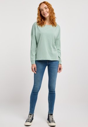 Giovane donna con capelli ricci rossi che sorride, indossa una maglietta a maniche lunghe verde chiaro, jeans skinny blu e sneakers nere alte.