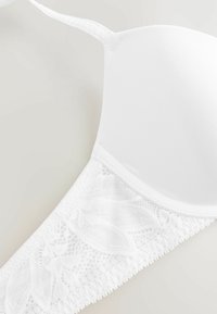 Witte beha met gladde cups en een kanten band. Verstelbare bandjes en een golvende afwerking voegen detail aan het ontwerp toe.