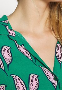 Camicia verde con motivi di foglie rosa bordati in blu navy. Colletto a scatto e tessuto testurizzato con una vestibilità rilassata. Un dettaglio a bottone è visibile.