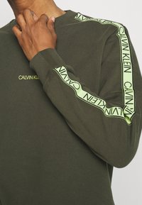 Sudadera de color verde oliva con un cuello y puños acanalados; presenta un logo destacado y franjas laterales de color verde lima en contraste con texto repetido.