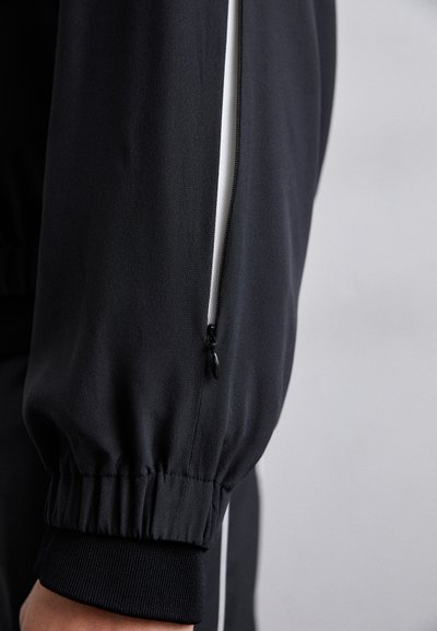Schwarze Sportjacke mit einem Reißverschlussbelüftungsfach, elastischen Bündchen und einem kontrastierenden weißen Seitenstreifen entlang des Ärmels.
