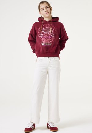 Sweat-shirt à capuche bourgogne avec un texte graphique, associé à un pantalon large blanc et des sneakers rouge et blanc. Tissu lisse, coupe décontractée.