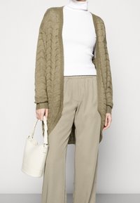 Olivgrüne Strickjacke mit texturiertem Muster, kombiniert mit einem weißen Rollkragenpullover. Das Modell hält eine cremefarbene Bucket Bag. Beigefarbene Weitbein-Hose.
