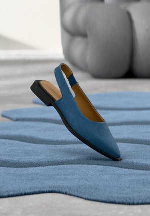 Zapato plano azul de tela con tira trasera, puntera puntiaguda y suela negra flotando sobre una alfombra azul esculpida con formas abstractas.