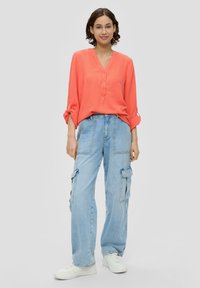 Korallenfarbenes Hemd mit V-Ausschnitt und hochgerollten Ärmeln; kombiniert mit hellblauen Cargo-Hosen mit großen Seitentaschen und weißen Sneakers.