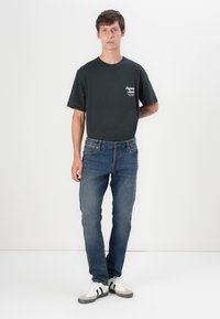 Camiseta de algodón negra con un gráfico blanco, combinada con jeans de mezclilla azul. El modelo lleva zapatillas blancas y grises con rayas negras.