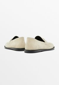 Beige suede loafers with a flat black sole, featuring a rounded toe and stitched detailing on the upper. Simple and minimal design.