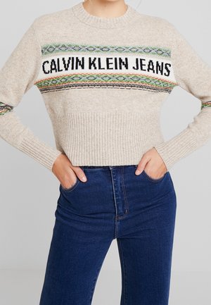 Maglione beige lavorato a maglia con la scritta "CALVIN KLEIN JEANS" e strisce decorative, abbinato a jeans blu scuro a vita alta, con le mani nelle tasche.