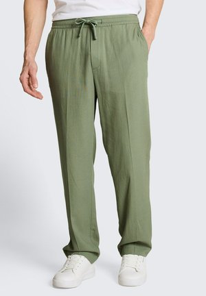 Mężczyzna noszący luźne oliwkowo-zielone spodnie z elastycznym pasem i troczkiem, połączone z białymi trampkami i białą koszulą.