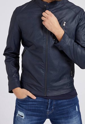 Man die een marineblauwe leren jas met ritssluiting en ritszakken draagt, met de ene hand in de zak en de andere hand de kraag verstelend.