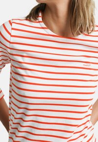 White cotton t-shirt with horizontal orange stripes, round neckline, and GANT logo in orange. Features three-quarter sleeves.
