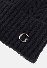 Fekete kötött sapka, bordázott textúrával és fonott kötés részletezéssel. Ezüst 'G' logóval ellátott fém díszítéssel.