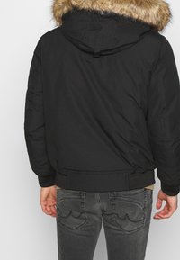 Schwarze Bomberjacke mit einem mit Pelz gefütterten Kapuze, die eine gesteppte Textur und gerippte Bündchen aufweist. Das Design auf der Rückseite hat eine glatte Oberfläche und einen verstellbaren Riemen.