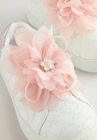 Valkoiset tennarit, joissa on teksturoitu muotoilu, vaaleanpunaisia kangaskukkia ja kultasävyinen "B" keskellä. Pehmeä materiaali ja koristeelliset yksityiskohdat.