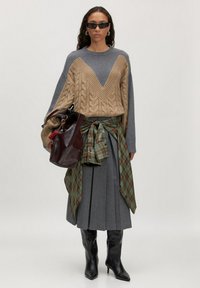 Beiger, grob gestrickter Pullover mit V-Ausschnitt, grauer, plissierter Rock, karierter Gürtel, braune Ledertasche und schwarze Stiefel mit Absatz. Die Accessoires umfassen eine Sonnenbrille.