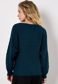 Maglione lavorato a maglia in teal con un pattern a trecce testurizzato, spalle svasate e polsini a coste. Design con schiena aperta che mette in risalto una vestibilità rilassata.
