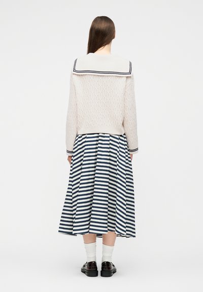 Kvinde stående med ryggen synlig, iført en cremefarvet cable-strikket sweater med en sømandsskrave og en navyblå og hvidstribet midinederdel.