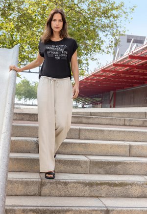 Zwarte t-shirt met witte tekst, gecombineerd met lichtbeige wijde broeken, gedragen met zwarte instappers, tentoongesteld op buitentrap.