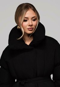 Mladá žena s blond vlasy upravenými do volných vln nosí velké zlaté kruhové náušnice a černou péřovou bundu s páskem a vysokým límcem.