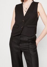 Gilet nero senza maniche su misura con due bottoni, pantaloni neri a vita alta dalla texture coordinata e collane a catena in argento sovrapposte.