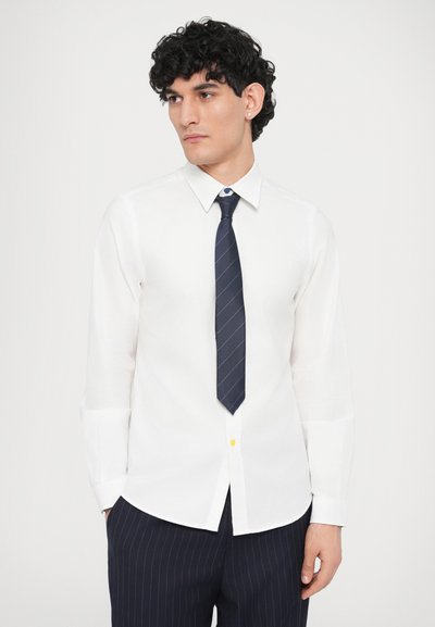 Camicia bianca a maniche lunghe con colletto, abbinata a una cravatta blu scuro con sottili strisce bianche. Piccolo pulsante giallo come dettaglio sulla camicia.