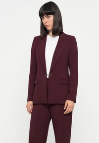 Bordeaux op maat gemaakte blazer met een gestructureerd ontwerp, enkelvoudige knoopsluiting en twee voorzakken, gecombineerd met een wit shirt.
