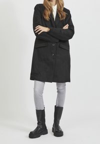Cappotto lungo nero con revers a punta, due tasche frontali e chiusura con bottoni, abbinato a jeans grigi slim-fit e stivali neri fino al ginocchio.