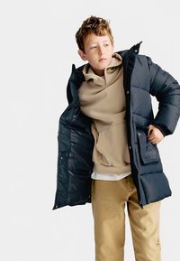 Dunkelblauer Steppmantel mit glänzender Textur, Reißverschlussverschluss und Seitentaschen, getragen über einem beigen Hoodie und einem cremefarbenen Hemd mit hellbrauner Hose.