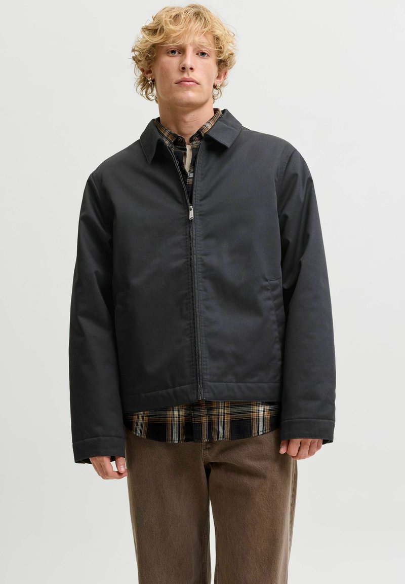 Jeune homme aux cheveux blonds bouclés portant une veste noire zippée par-dessus une chemise à carreaux et un pantalon marron, debout devant un fond uni.