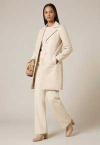 Femme portant un long manteau beige, un pantalon à jambes larges et des bottines, tenant un sac à main beige assorti sur un fond neutre uni.