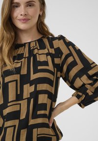 Black and gold patterned blouse with a loose fit, gathered neckline, and voluminous sleeves featuring elastic cuffs. Textured fabric with geometric design.