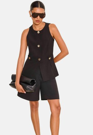 Frau trägt ärmellosen schwarzen Blazer mit goldenen Knöpfen, schwarze Shorts, dunkle Sonnenbrille und hält eine große schwarze Clutch.
