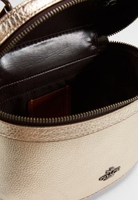 Okrąła, metaliczna złota torebka skórzana o teksturowanym wykończeniu. Wnętrze z ciemnobrązowej tkaniny z małą kieszonką na karty. Zamek błyskawiczny.