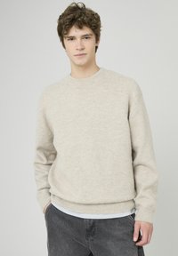 Pull en maille beige clair avec un col rond, poignets et ourlet côtelés, porté sur une chemise blanche. Texture douce, coupe décontractée.
