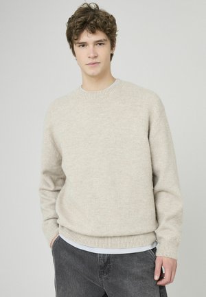 Pull en maille beige clair avec un col rond, poignets et ourlet côtelés, porté sur une chemise blanche. Texture douce, coupe décontractée.