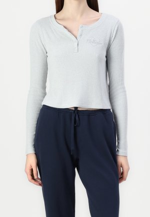 Femme portant un haut court côtelé gris clair à manches longues avec des boutons et un pantalon bleu marine à cordon, visible du cou aux hanches.