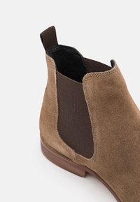 Bottine Chelsea en daim beige avec panneau latéral élastique sombre, doublure intérieure noire, languette de tirage au talon et semelle en bois sur fond blanc.