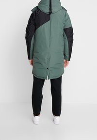 Vadderad jacka i grön och svart, med en huva, kontrasterande paneler och en något längre bakkant. Bärs med svarta byxor och vita skor.