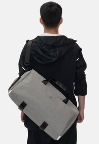 Grå tygtrékkväska med svarta läderdetaljer och -handtag, rektangulär form och logotypdetalj på sidan. Bärs över axeln.