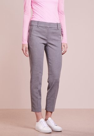 Person trägt graue Slim-Fit-Hosen, ein pinkfarbenes Langarmoberteil und weiße Turnschuhe, steht auf einem beigen Boden vor einem hellrosa Hintergrund.