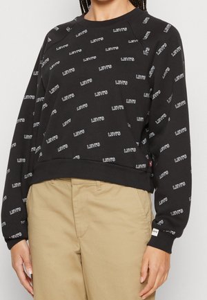 Sort langærmet sweatshirt med gentagne hvide "LEVI'S"-logoer, kombineret med beige bukser, vist fra midt på torsoen til halsen.