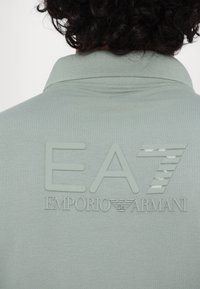 Šviesiai žalia polo maikutė su tekstūriniu "EA7" logotipu ir balta spalva atspausdintu "EMPORIO ARMANI", su klasikiniu apykaklės dizainu ir minkšta medžiaga.