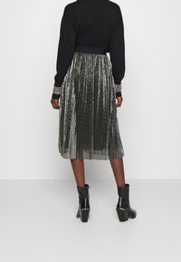 Falda midi plisada metálica en verde oscuro con un acabado brillante. Combinada con un suéter negro con puños adornados y botas de tobillo.