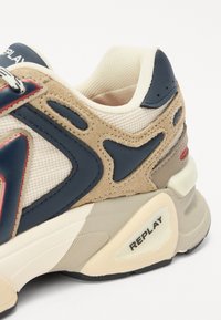 Beige en marineblauwe sneaker van mesh en suede materialen, met rode accenten, een ronde hak en een zichtbare merklabel op de zool.