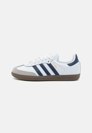 Hvid Adidas Samba-sneaker med marineblå striber, gummisål og grå ruskindståkappe, vist fra siden på en ensfarvet baggrund.