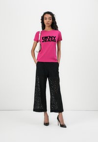 Fuksia t-paita, jossa on "DKNY JEANS" logo, yhdistettynä löysiin mustiin pitsihousuihin ja mustiin korkokenkiin. Valkoinen käsilaukku asusteena.