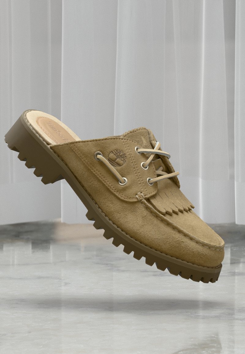 Mocassin à enfiler en daim beige avec semelle robuste, détails de lacets, franges à l'avant et logo embossé sur le côté, présenté sur un fond gris et blanc.