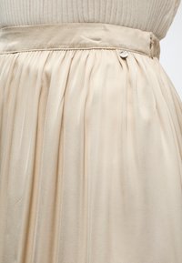 Μπεζ πλισέ φούστα με ψηλή ζώνη και μικρή μεταλλική λεπτομέρεια κουμπιού, φορεμένη με ένα ελαφρύ ριμπ πουλόβερ.